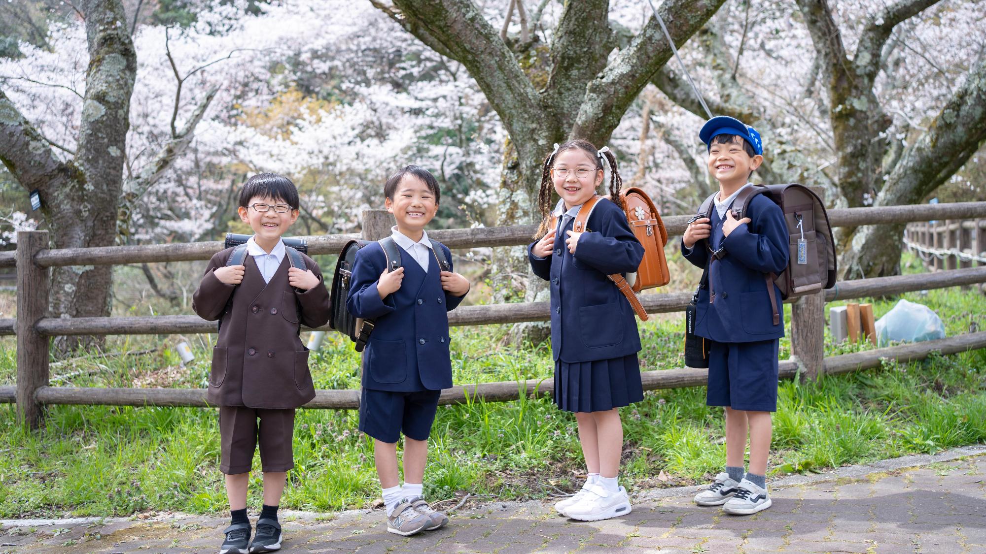 4人の子どもが桜の下でランドセルを背負って笑顔を向けている写真