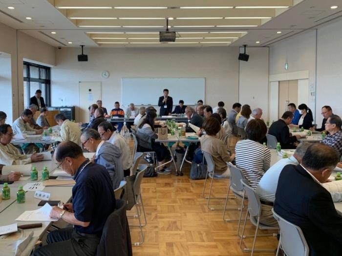 会議室内の奥に設置されたホワイトボードの前で男性がマイクを片手に話しをしているのを、参加者が席について話しを聞いている写真