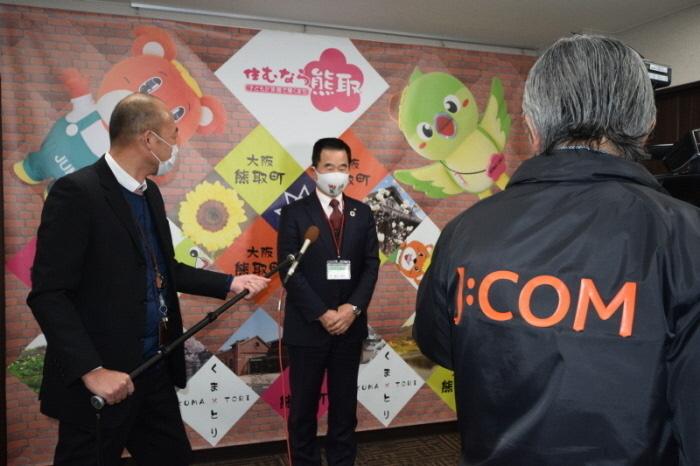 町長がスーツ姿の男性から差し出されたマイクで話をしていて、ビデオカメラで撮影をされている写真