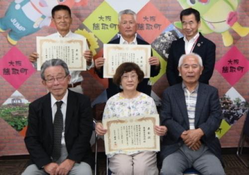 中央に表彰状を持った女性、その両脇に表彰状を持った男性2人がいすに腰掛け、後ろに町長と表彰状を持った男性2人が並んで立っている記念写真