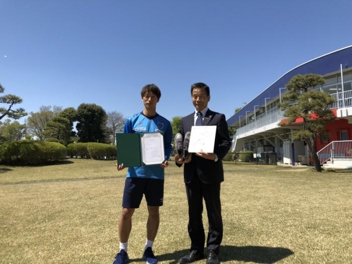 芝生のグラウンド上で任命書を持った室屋選手とサイン色紙を持った町長が並んで立っている記念写真