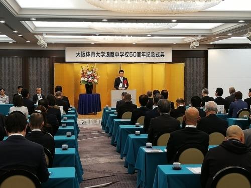 金の屏風がある舞台で町長が話をしていて、スーツを着た出席者たちが座って話を聞いている写真