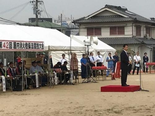広場に置かれた赤い台の上で町長が話をしていて、奥にあるテントの下で消防団の人たちが座って話を聞いている写真