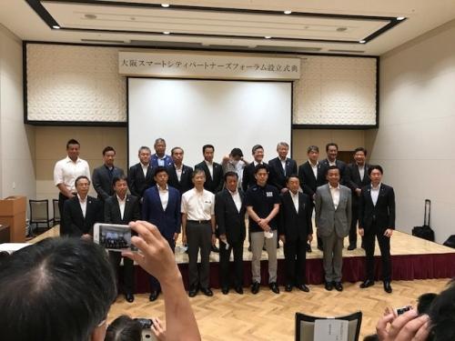 前方の舞台に20名ほどのスーツ姿の男性が前後2列に並んでいる集合写真
