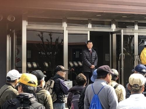 建物の入口の前の帽子を被ったり、リュックを背負っている参加者たち、入口側のガラスの扉の前に立っている町長が写っている写真