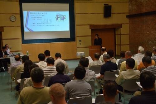 大きなスクリーンを見ながら演台の前で話す町長と、座って聞く参加者の写真