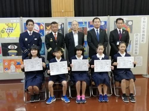 奥には絵画作品が飾られていて、手前には5名の賞状を持った小学生たちとスーツ姿の5名の男性たちが写っている写真