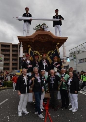 黒い法被を着た男性たちが神輿の前に並んでいて、神輿の上にも2人男性が立っている写真