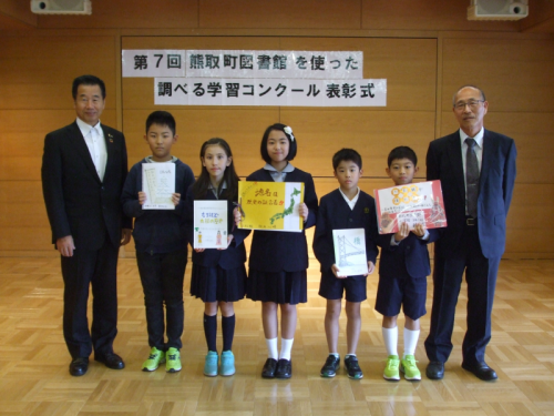 絵などが描かれた作品を持った5名の小学生たちと町長、スーツ姿の男性が並んでいる記念写真