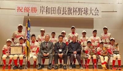 ユニホームを着て、首にメダルを掛けた熊取ジャガーズの皆さんが優勝旗とトロフィー、縦に入った賞状を持って、町長と関係者の方と一緒に2列に並んで写っている集合写真