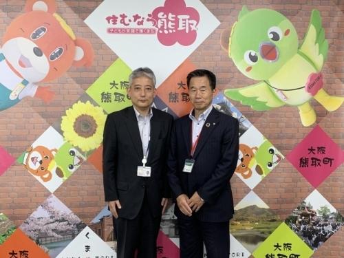 ゆるキャラが描かれた背景の前で並んで立つ町長と佐伯亮介さんの写真