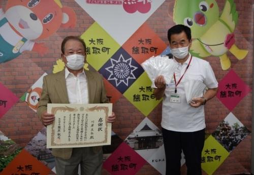 個包装の白いマスクを両手に沢山持っている町長と感謝状を持っている川岸さんの写真