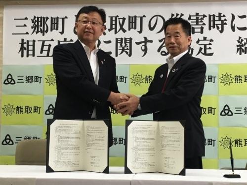 机の上に協定書を置き、その後ろで両手で握手をする町長と三郷町関係者の写真
