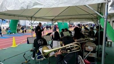 白いテントの下に座って大阪観光大吹奏楽部が演奏しているくまとりロードレース会場の様子の写真