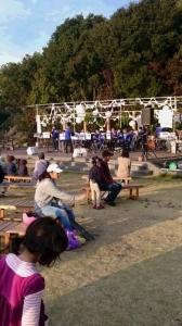 ワンダーフォレストの屋外ステージ上の大阪観光大吹奏楽部の演奏を、木製の椅子に座っている観客が聞いている写真