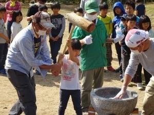 大勢の子ども達が見守るなか、白いシャツを着た女の子が男性スタッフと一緒に餅つきを行っている様子の写真