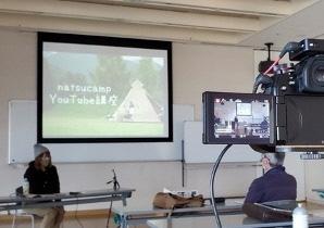 スクリーンに映し出された映像の前にニット帽を被った女性が座り、向かい合って男性が座っているYouTuber養成講座の様子の写真