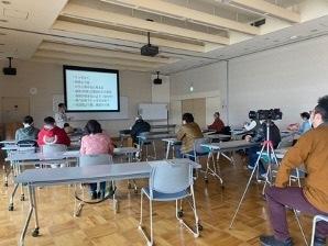教室の前方に設置されたスクリーンの左側に講師が立ち、席に座った参加者がスクリーンに映し出された資料を見ているYouTuber養成講座の様子の写真
