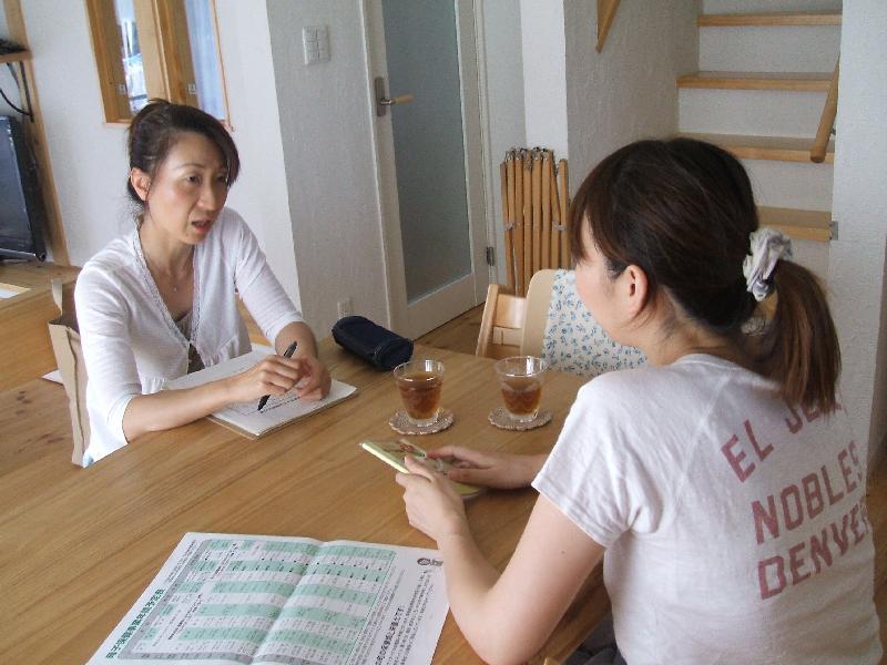 訪問宅のテーブルで、助産師の女性と母子手帳を持っている女性が向い合って座っており、話をしている助産師訪問の様子の写真