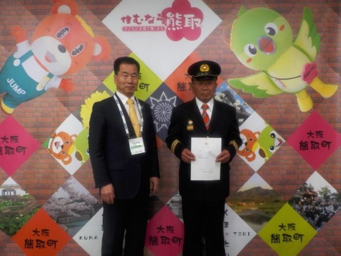 「住むなら熊取」と書かれた背景の前で、町長と新消防団長の辞令を両手で持った相輪氏が横に並んでいる写真