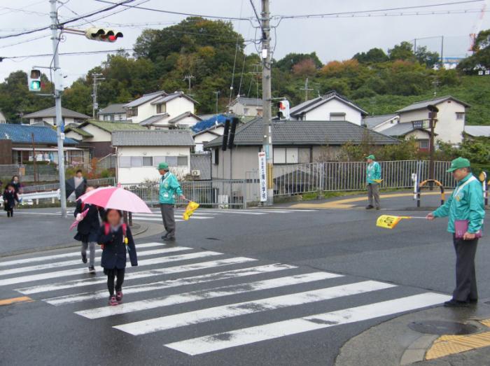 傘をさして横断歩道を渡る子供たちを見守っている、緑色のキャップにジャケットを着た見守り活動隊の写真