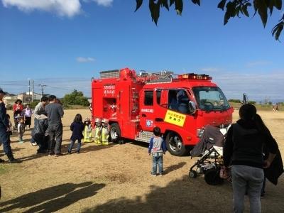 広場に停まった消防車の周りに子ども達が集まり消防士体験を行っている写真