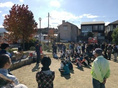 屋外広場で消防職員による説明を参加者が地面に座ったり立ったりして聞いている写真