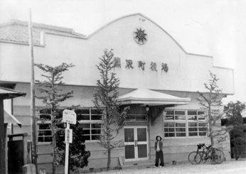 熊取町役場の建物の前に一人の女性が立っていて、横には自転車、木などがあるモノクロ写真
