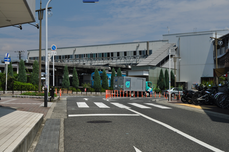 奥には横長の鉄筋で造られた駅が立っていて、手前にはアスファルトの道路が写っているカラー写真