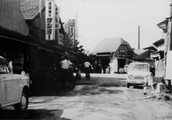 駅の前のお店などがある通りで人が歩いていたり車が通っている様子が写っているモノクロ写真