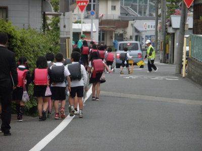歩道を歩いている小学生と交差点に立って子供を誘導している子ども見まもり隊の写真