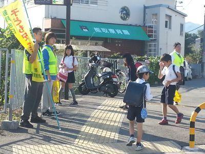 道路の脇に立っている学生版子ども見まもり隊とランドセルを背負った小学生の写真