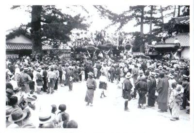 奥には大きな神輿と神輿の上に立っている人たちが写っていて、手前にはカンカン帽をかぶり浴衣を着た人たちが集まっているモノクロ写真