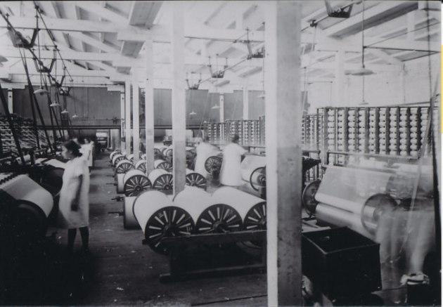 大きな丸いシャットルや自動織機が並び、白い服を着た複数の女性がそれぞれ織機を操作している中林綿布工場の様子