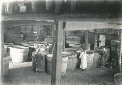 義本酒造の発酵場内にて、所狭しと並ぶ大きな樽とその前に立つ3人の青年の白黒写真