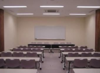 椅子が3脚ずつ付けられた複数の長机と、その正面にホワイトボードがある講義室の写真