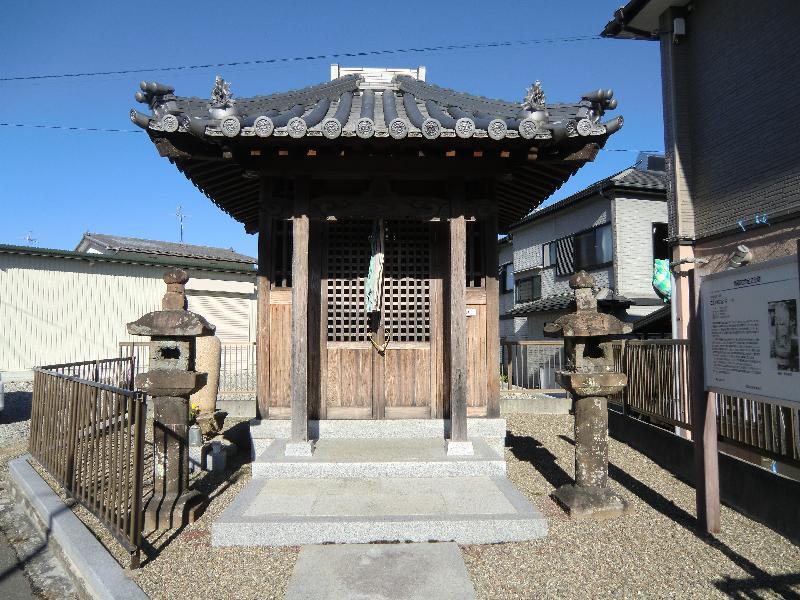 中央に石造地蔵菩薩立像が祭られた祠が建っており、その両側に石灯籠と右側に案内看板が立っている写真