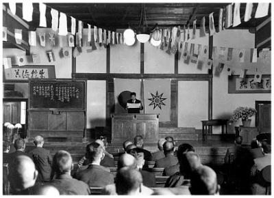 小学校の講堂内にて、壇上で講話をする男性と、椅子に座り熱心に話を聞く参加者達の白黒写真