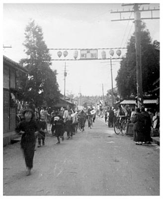 電柱程の高さに設置された、祝町制と書かれた紅白の門と、その下の道路を行き交う人々の白黒写真
