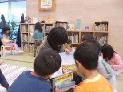 アトム共同保育園の室内で、女性が絵本の読み聞かせを行っているのを近くに来て熱心に聞いている子供たちの写真