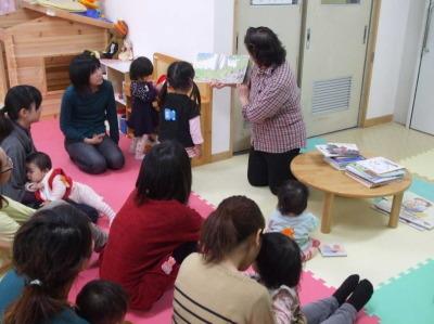 教育子どもセンターの室内で、女性が絵本を持って読み聞かせを行ってのを、親子で参加している子供たちが聞き入っている写真