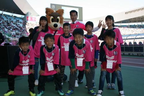 ピンクのユニフォーム姿の子供たち8人と町長、セレッソ大阪のマスコットキャラクターの着ぐるみが並んでいる記念写真