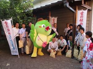 メジーナちゃんと、タスキを付けた関係者や町長の集合写真
