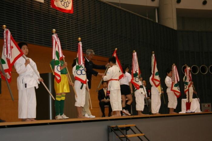 舞台上で空手着やサッカーのユニフォーム姿の子供たちが旗を持ち横並びに並んでいる写真