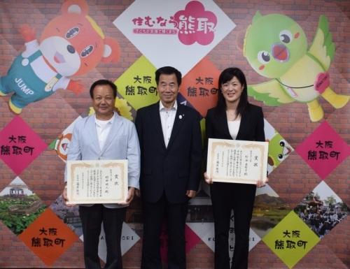 ジャンプ君とメジーナちゃんのが描かれた壁の前で左から村田明人さん、町長、杉谷美智子さんが並んでいる記念写真