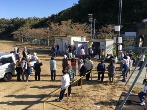 広場の奥に防災倉庫、倉庫の前には消防団員、手前には参加者達やトラックが写っている取扱訓練の写真