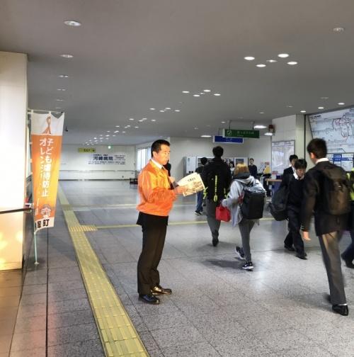オレンジの上着を着た町長が、駅の構内を歩いている人たちに広報誌を配ろうとしている写真