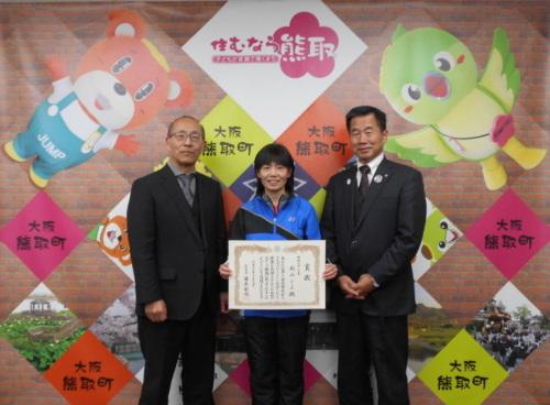 ジャンプ君とメジーナちゃんのが描かれた壁の前で、左から教育長、秋山とよさん、町長が並んでいる記念写真