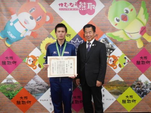 ジャンプ君とメジーナちゃんのが描かれた壁の前で、賞状を持った稲葉潤也さんと町長が並んでいる記念写真