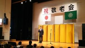 祝敬老会と書かれた幕と金屛風が置かれた舞台上でスーツを着た町長が話をしている写真